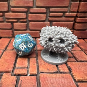 May include: A grey, spiky, tree-like miniature figure and a blue and green 20-sided die. The die has white numbers and is next to the miniature on a brick-patterned surface. The miniature is on a round base.
