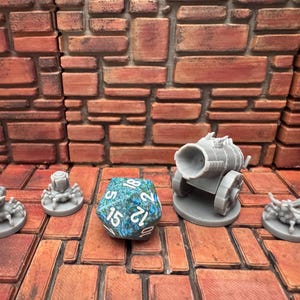 May include: A collection of miniature gray fantasy game pieces, including a cannon, spiders, and a multi-sided die. The die is teal and blue with white numbers. The pieces are arranged on a brick-patterned surface.