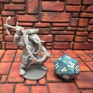 May include: A gray miniature figure of an elephant-headed character holding a chain and a weapon, next to a blue and green twenty-sided die. The figure is on a round base, set against a brick wall.