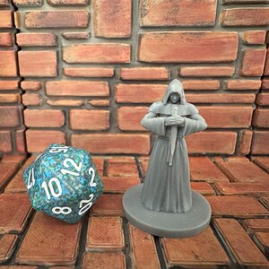 May include: A gray painted miniature figurine of a hooded figure holding a dagger, standing on a gray base. The figurine is in front of a brick wall and a blue and green dice.