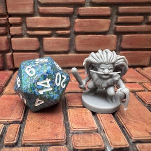 May include: A blue and green 20-sided die with white numbers sits beside a grey miniature figure of a fantasy character holding a club and a weapon. The items are on a brick-patterned surface.