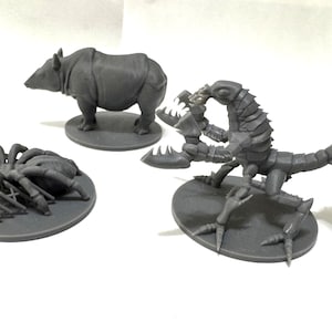 May include: Three unpainted grey resin miniature figures on circular bases. One figure is a rhinoceros, one is a large spider with multiple legs, and the third is a creature with a segmented body, large pincers, and a long, spiked tail.