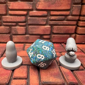 May include: A selection of tabletop gaming accessories. Features a 20-sided die with blue and green patterns, and two grey miniature figures. The background has a brick-like texture.