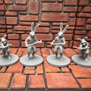 May include: Four gray miniature figurines of anthropomorphic rabbits. Two stand upright holding a staff, while the other two are crouched, holding a bow and arrow. The figures are on gray circular bases, set against a brick wall.