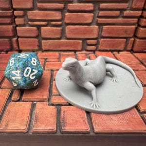 May include: A grey lizard figurine on a grey circular base, beside a blue and green twenty-sided die. The objects are on a brick-patterned surface, with a brick wall in the background. The die has white numbers.