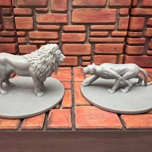 May include: Two gray lion and panther figurines on circular bases. The lion stands upright, while the panther is in a stalking pose. The background features a brick wall and a wooden surface.