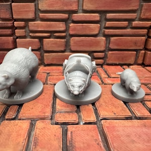 May include: Three gray raccoon figurines of varying sizes. One raccoon is standing, another is wearing a helmet, and the smallest is in a walking pose. Each figurine is on a circular base. The background features a brick wall and floor.