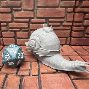 May include: A gray, 3D-printed snail figurine wearing a helmet and armor, next to a blue and green twenty-sided die. The snail is on a brown surface with a brick wall background. The snail's shell has a detailed texture.