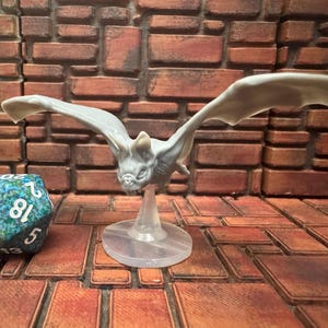 May include: A gray bat figurine with outstretched wings, mounted on a clear stand. A multi-sided die with blue and green accents is to the left of the bat. The background is a brick wall.