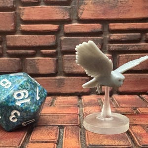 以下が含まれることがあります： 青と緑の大理石模様のD20サイコロと、小さな薄い灰色のフクロウの置物が飛んでいる様子。 フクロウは透明なスタンドに取り付けられています。 背景はレンガの壁です。
