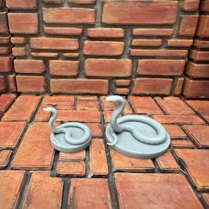 May include: Two gray snake figurines, one small and one larger, are displayed on a textured, brick-like surface. The snakes are coiled with their heads raised, showcasing a detailed design. The background features a similar brick pattern.