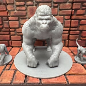 May include: A gray gorilla figurine stands on a gray circular base, flanked by two smaller gorilla figurines. The figurines are set against a brick background, showcasing a detailed primate sculpture.