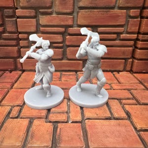 Può includere: Due figurine grigie in miniatura di guerrieri, ognuno con una grande ascia. Le figure sono su basi circolari, in posa come se fossero in azione. Lo sfondo è un motivo a forma di mattone nei toni del marrone e dell'arancione, che suggerisce un tema di gioco di ruolo.