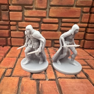 May include: Two gray miniature figures of hooded characters, each holding a sword. They are posed in a dynamic stance, with cloaks and detailed armor. The figures are on round bases, suitable for tabletop gaming.