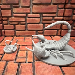 May include: Two gray scorpion figurines, one large and one small, are displayed on a textured, orange surface. The larger scorpion is on a circular base, with detailed claws and a curved tail. The background features a brick wall.