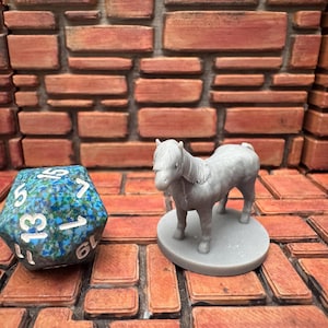 May include: A gray miniature pony figurine on a round base stands next to a blue and green polyhedral die. The die has white numbers. The background is a brick wall.