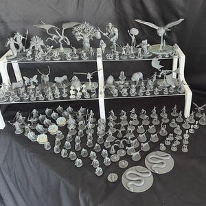 May include: A collection of gray, 3D printed miniature figures displayed on a two-tiered shelf. The figures include a variety of creatures, people, and objects, such as a dragon, a tree, a bird, and a snake.