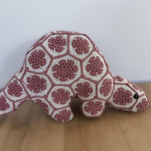 May include: A handmade crochet dinosaur plushie in dusty rose and white. The dinosaur has a geometric pattern and a button eye. The plush toy is lying on a wooden surface.