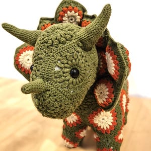 Può includere: Un dinosauro Triceratops all'uncinetto fatto a mano, nei toni del verde, arancione e bianco. Il dinosauro presenta un design dettagliato e strutturato con corna, un collare e occhi neri a bottone. Il lavoro all'uncinetto è intricato, con un motivo floreale.