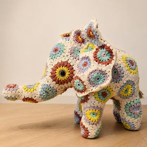 Könnte beinhalten: Ein Stoffelefant, der aus zahlreichen gehäkelten Granny Squares in einer cremefarbenen Grundfarbe mit Akzenten in Gelb, Hellblau, Lavendel und Rostorange gefertigt ist. Der Elefant hat einen Rüssel, große Ohren und ein dunkles, kreisförmiges Auge.