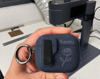 May include: A dark blue textured protective case for wireless earbuds featuring a black rectangular clip, a silver metal keyring, and a laser-engraved design of a single rose with the name Naomi printed in a white serif font on the front surface.