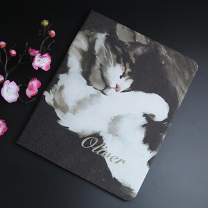 May include: A notebook with a watercolor painting of a cat. The cat is white and gray, with the name "Oliver" in cursive script. The background is dark gray. Pink flowers are in the upper left corner.