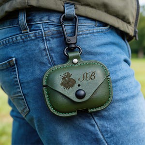 Op de afbeelding: Een donkergroene leren AirPod-hoes met een zwarte drukknoopsluiting en een karabijnhaak. De hoes heeft een reli&euml;fontwerp van een figuur en de initialen "SB". De hoes is bevestigd aan een spijkerbroek.