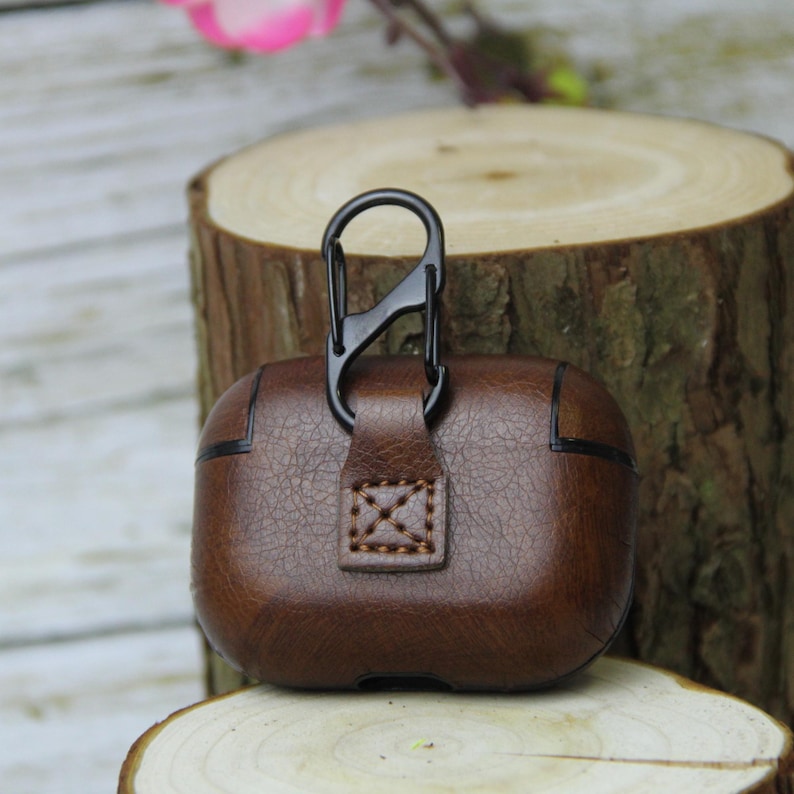May include: Brown leather earbud case with a black metal carabiner clip. The case has a textured finish and a stitched accent. The case is designed to protect earbuds and is shown on a wooden surface.