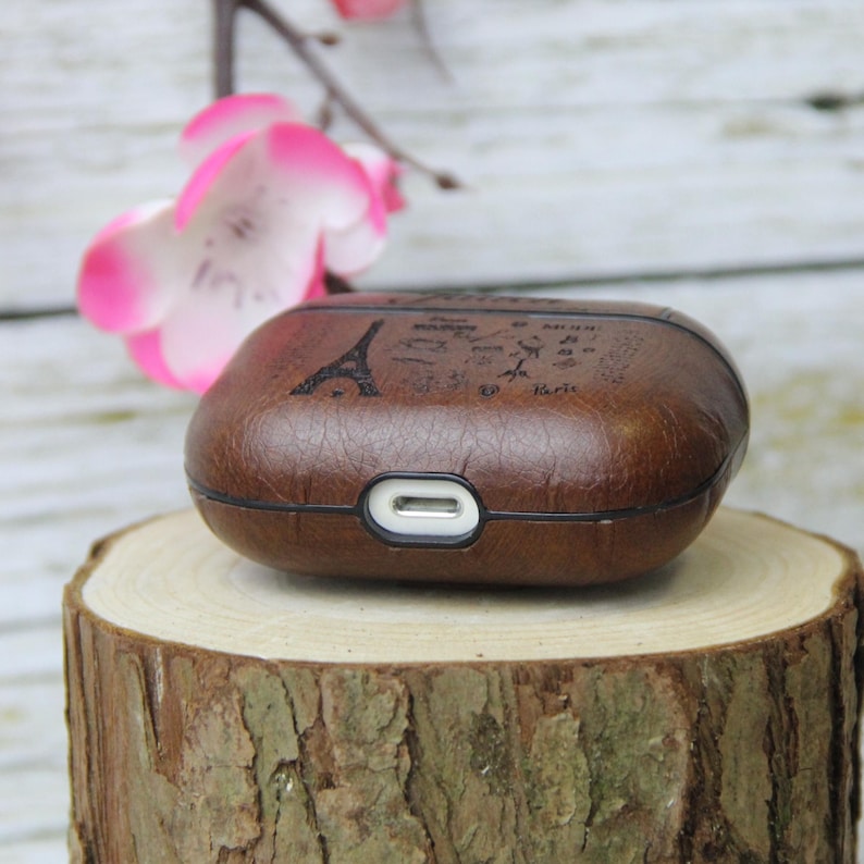 May include: Brown leather AirPods case with a design featuring the Eiffel Tower and the word "Paris". The case has a charging port at the bottom and is sitting on a wooden surface. Pink flowers are in the background.