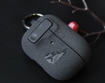 May include: A dark gray textured protective case for wireless earbuds featuring a matte finish, a black carabiner attachment, and the name Naomi engraved in script. The case displays a small etched graphic of a whale tail above the name.