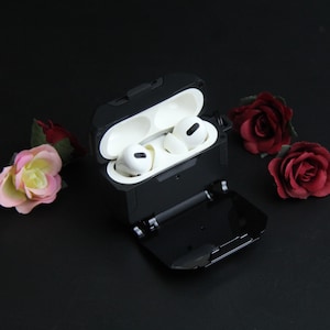 Puede incluir: Una funda protectora negra para auriculares inal&aacute;mbricos, abierta para revelar auriculares blancos en el interior. La funda es rectangular con una tapa con bisagras. Rosas rojas y rosas est&aacute;n dispuestas alrededor de la funda sobre una superficie negra.