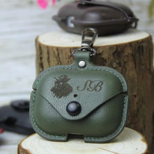 Op de afbeelding: Groene leren AirPod-hoes met een klepsluiting en een zilverkleurige karabijnhaak. De hoes heeft een gegraveerd ontwerp en de initialen "SB". Een tweede, vergelijkbare hoes is zichtbaar op de achtergrond.