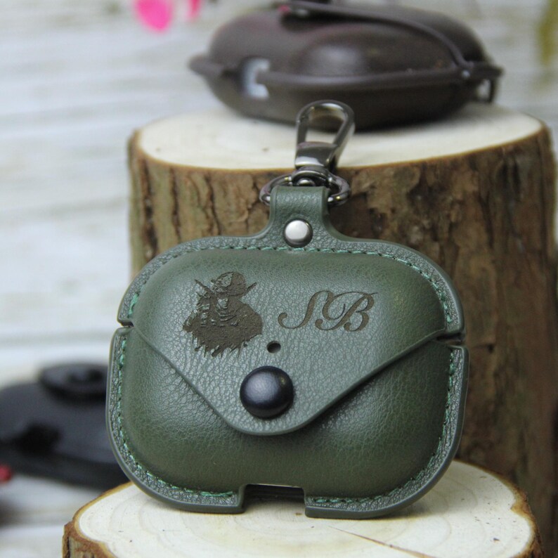Op de afbeelding: Groene leren AirPod-hoes met een klepsluiting en een zilverkleurige karabijnhaak. De hoes heeft een gegraveerd ontwerp en de initialen "SB". Een tweede, vergelijkbare hoes is zichtbaar op de achtergrond.