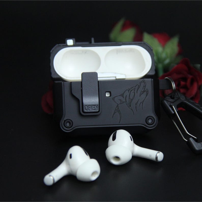 May include: Black protective case for wireless earbuds featuring a wolf howling design. The case is open, showing the interior, and has a carabiner clip. Two white earbuds are in front of the case.