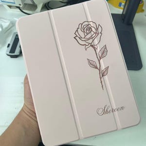 Może przedstawiać: Jasnoróżowe etui na tablet z ciemnobrązową grafiką liniową przedstawiającą pojedynczą różę z liśćmi i łodygą. Imię Shereen jest nadrukowane kursywą w prawym dolnym rogu przedniej części składanego etui.