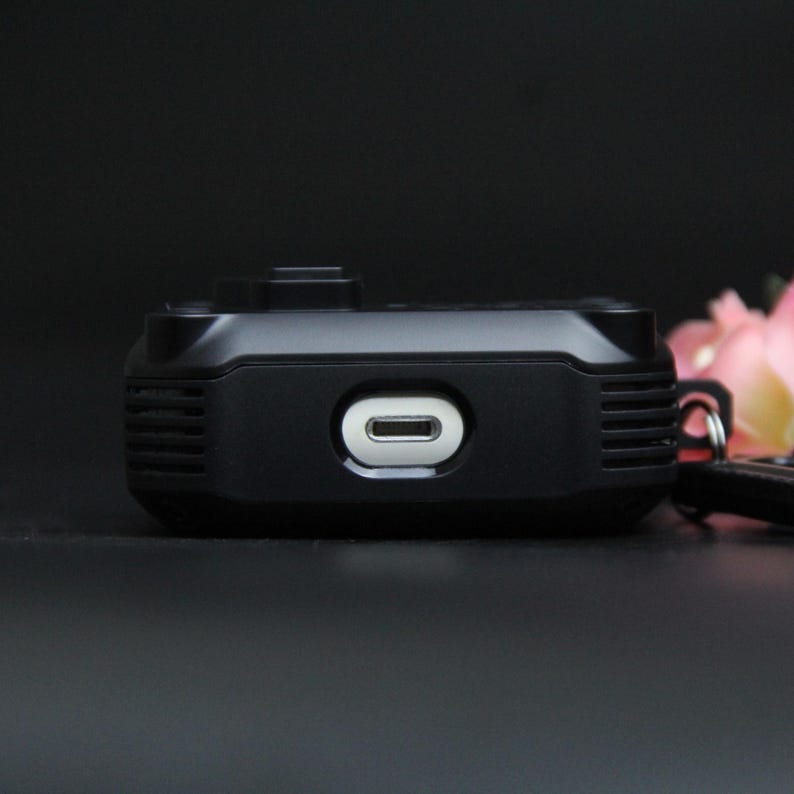 May include: A small, black rectangular device with a white oval port. The device has a matte finish and a ridged texture on the sides. The background is dark, with a hint of pink flowers.