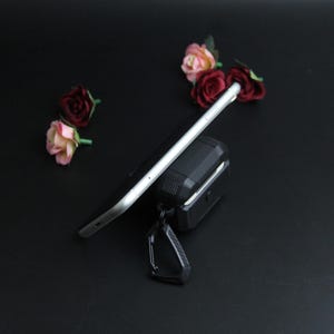Puede incluir: Un tel&eacute;fono inteligente negro descansa sobre una funda protectora negra con un mosquet&oacute;n. El tel&eacute;fono es plateado y negro. Rosas artificiales rosas y rojas est&aacute;n esparcidas sobre la superficie negra.