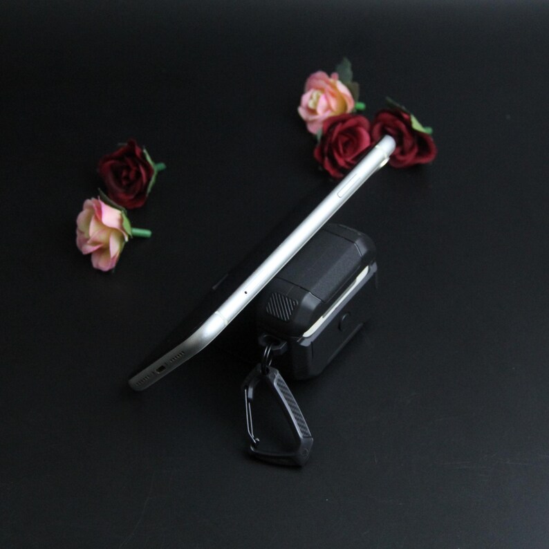 Puede incluir: Un tel&eacute;fono inteligente negro descansa sobre una funda protectora negra con un mosquet&oacute;n. El tel&eacute;fono es plateado y negro. Rosas artificiales rosas y rojas est&aacute;n esparcidas sobre la superficie negra.