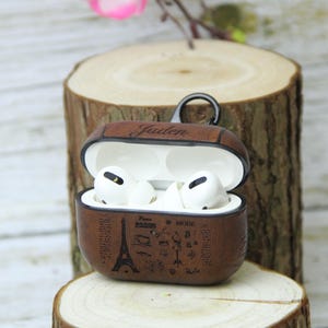 May include: Brown leather AirPods Pro case with a black metal clasp, open to reveal white earbuds. The case is decorated with a Paris-themed design, including the Eiffel Tower and the word "Paris". The name "Jaiden" is embossed on the top.