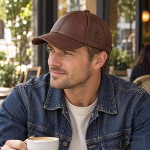 Può includere: Un cappellino da baseball in similpelle marrone con un design strutturato a sei pannelli e visiera curva. Il lato del cappellino presenta una piccola grafica in rilievo di una rosa sopra il testo NAOMI in un carattere sans-serif pulito.