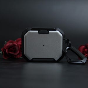 Puede incluir: Funda protectora para auriculares con un panel central plateado y un marco negro resistente. Incluye un mosquet&oacute;n para sujetar. Rosas rojas en el fondo.