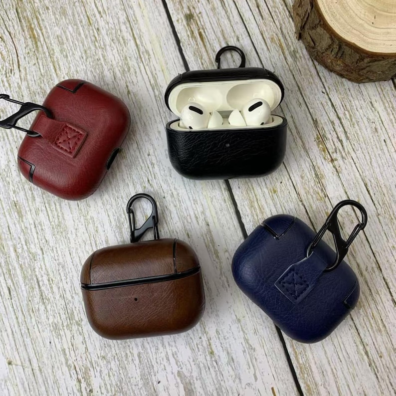 May include: Four leather AirPod Pro cases in red, black, brown, and blue. The black case is open, revealing white earbuds. Each case has a black carabiner clip and a textured finish.