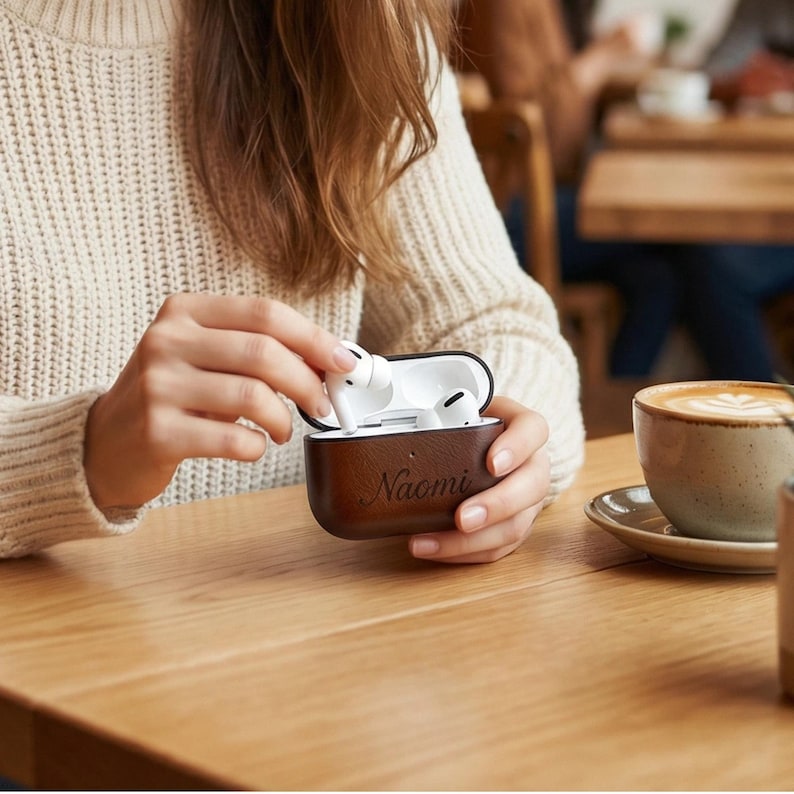 Peut inclure: Un &eacute;tui en cuir marron pour AirPods avec le nom "Naomi" grav&eacute;. L'&eacute;tui est ouvert, r&eacute;v&eacute;lant des AirPods blancs. Une tasse de caf&eacute; sur une soucoupe est pos&eacute;e sur une table en bois.