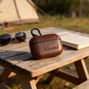 Peut inclure: Un &eacute;tui en cuir marron pour &eacute;couteurs sans fil, grav&eacute; du nom "Naomi". L'&eacute;tui est pos&eacute; sur une table en bois, avec des lunettes de soleil et un livre en arri&egrave;re-plan. Il poss&egrave;de une boucle noire pour l'attacher.