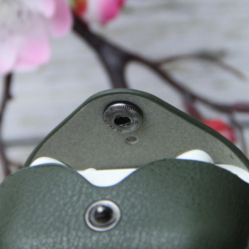 Op de afbeelding: Close-up van een donkergroene leren etui met een druksluiting. De etui is open en onthult een lichtgrijze binnenkant en witte rand. Een metalen drukknop is zichtbaar aan de bovenkant en een andere aan de onderkant. De achtergrond is vervaagd met roze en bruine tinten.