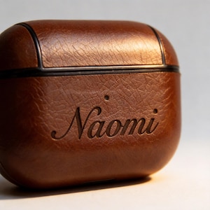 Peut inclure: Un &eacute;tui en cuir marron pour AirPod avec le nom "Naomi" grav&eacute; sur le devant. L'&eacute;tui a une surface textur&eacute;e et une bordure noire sur le dessus. L'&eacute;tui est con&ccedil;u pour prot&eacute;ger et transporter des &eacute;couteurs sans fil.