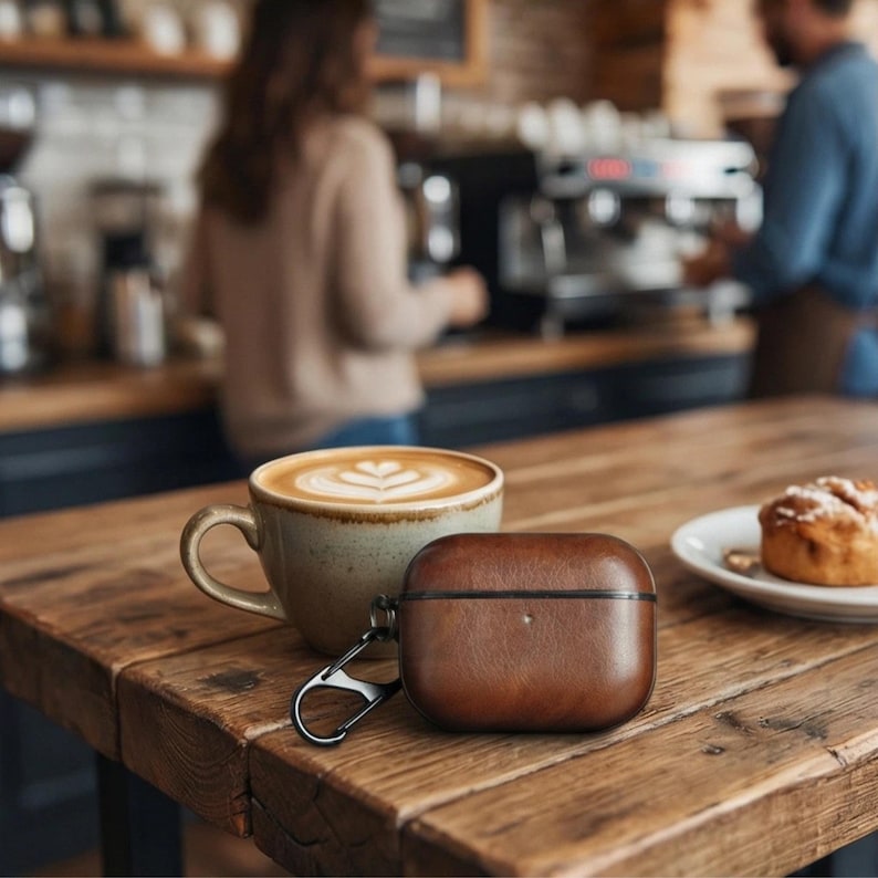 Peut inclure: Un &eacute;tui en cuir marron pour AirPods avec un mousqueton argent&eacute; est pos&eacute; sur une table en bois &agrave; c&ocirc;t&eacute; d'un caf&eacute; au lait et d'une p&acirc;tisserie. L'&eacute;tui est rectangulaire et a une bordure marron fonc&eacute;. La tasse &agrave; caf&eacute; est en c&eacute;ramique et de couleur marron clair.