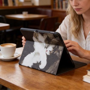 Puede incluir: Funda negra para tableta con una ilustración de un gato blanco. La funda está abierta, mostrando la pantalla. Una taza de café está sobre la mesa de madera. La ilustración del gato tiene acentos grises y marrones.