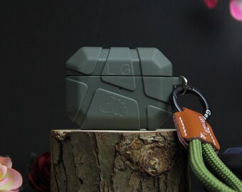 May include: An olive green earbud case with a geometric pattern and a small embossed graphic. It's attached to a green and brown lanyard with a leather tag. The case is displayed on a wooden block. The case is designed to protect the earbuds.