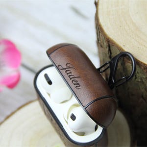 May include: Brown leather AirPod case with the name "Jaden" engraved on the lid. The case is open, revealing white earbuds inside. A black carabiner clip is attached to the side. The case is on a wooden surface.
